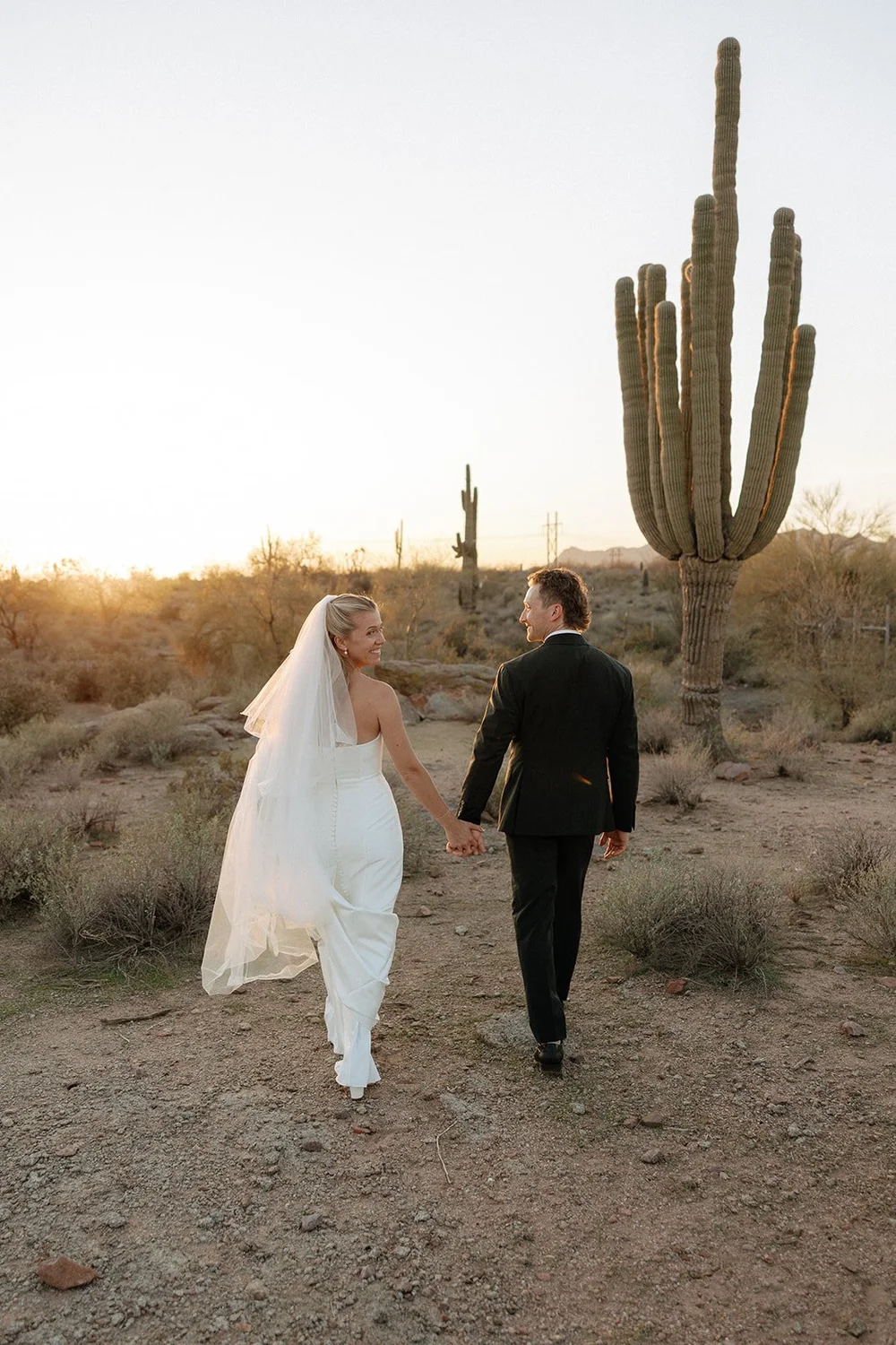 A bride and groom embrace in wedding attire in a desert landscape, with a tall cactus in the background