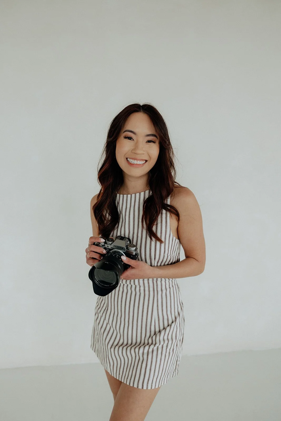 A woman in a striped sleeveless dress holds a camera and smiles, standing against a plain white background.