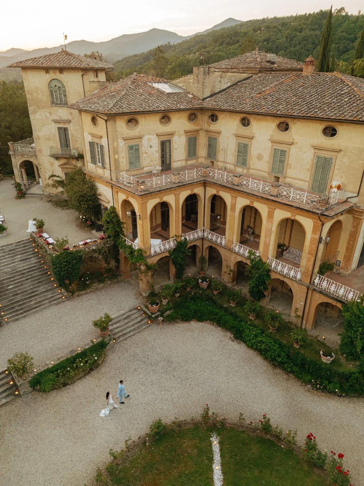 Aerial view of a large historic villa with balconies, stone steps, and a gravel courtyard surrounded by greenery and hills. Two people are walking near the building for an italy destination wedding