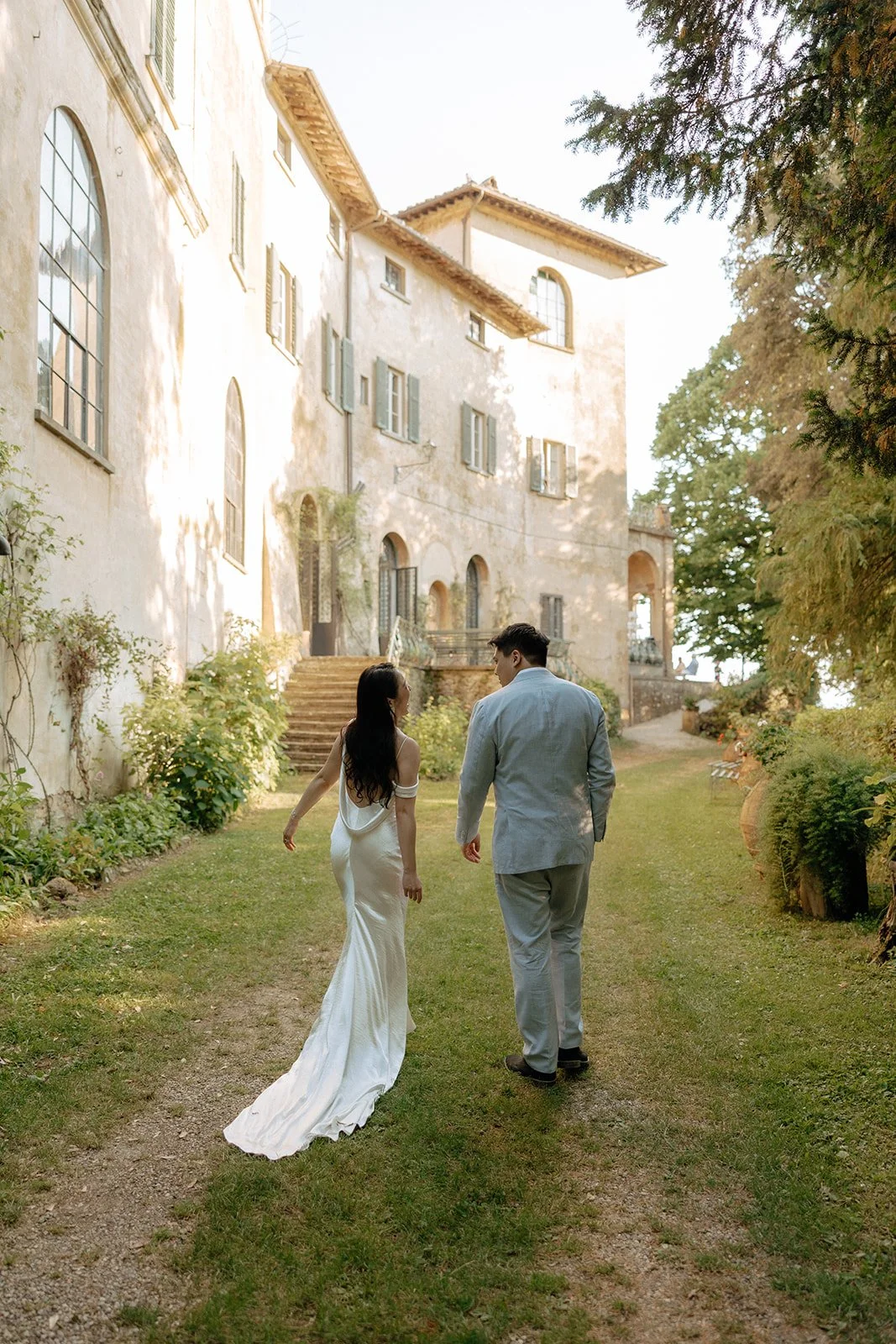 Aerial view of a large historic villa with balconies, stone steps, and a gravel courtyard surrounded by greenery and hills. Two people are walking near the building for an italy destination wedding