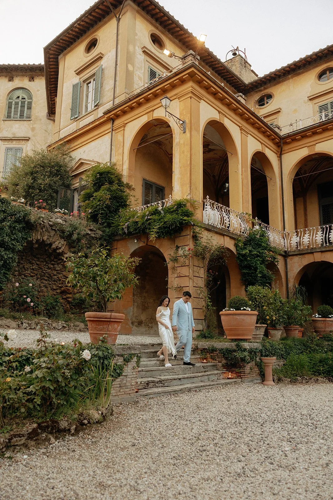 Aerial view of a large historic villa with balconies, stone steps, and a gravel courtyard surrounded by greenery and hills. Two people are walking near the building for an italy destination wedding