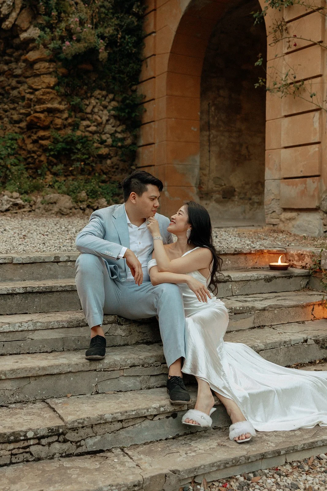 A couple in formal attire sits on stone steps outdoors, looking at each other. The woman wears a white dress, and the man is in a light-colored suit.