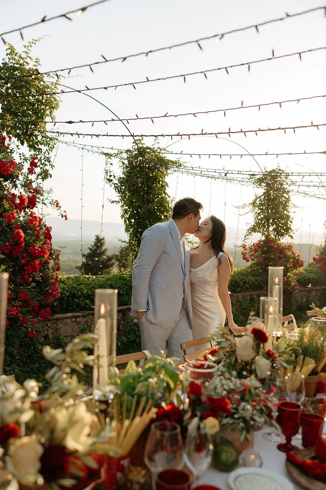 A couple dressed in formal attire kisses in an outdoor setting, surrounded by flowers, greenery, string lights, and a table set for a meal.