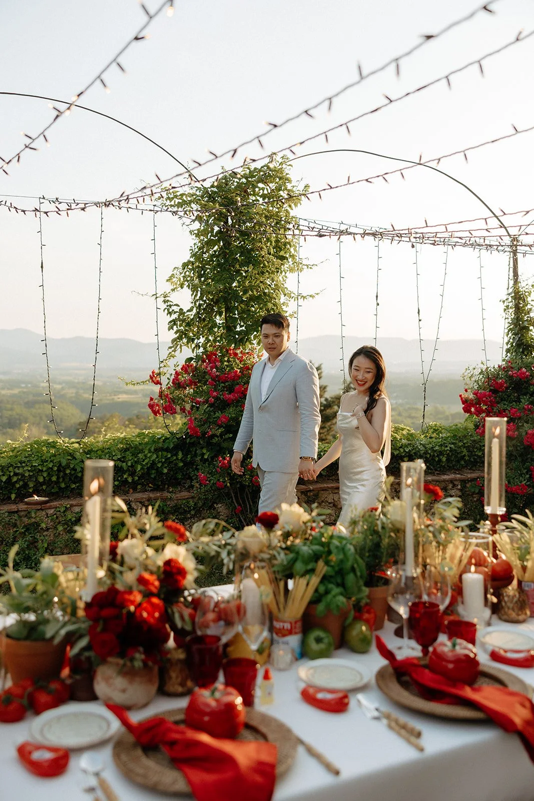 A couple dressed in formal attire kisses in an outdoor setting, surrounded by flowers, greenery, string lights, and a table set for a meal.