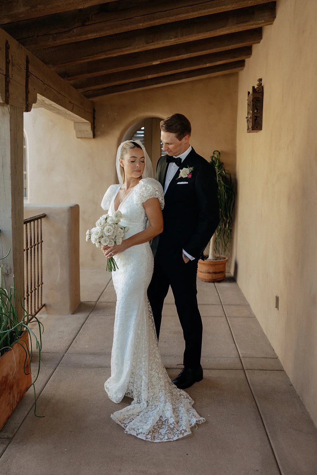 A bride in a white gown holding a bouquet stands next to a groom in a black suit and bow tie in a corridor with tan walls and wooden beams for a destination wedding in Italy