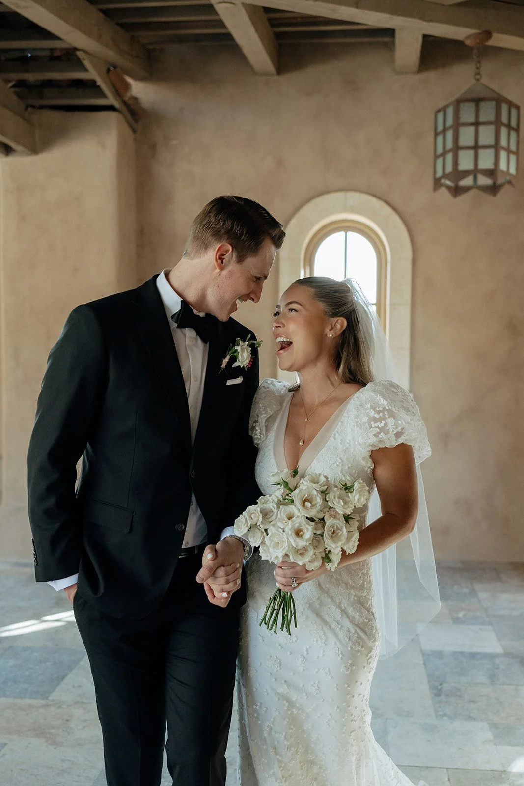 A bride in a white gown holding a bouquet stands next to a groom in a black suit and bow tie in a corridor with tan walls and wooden beams for a destination wedding in Italy
