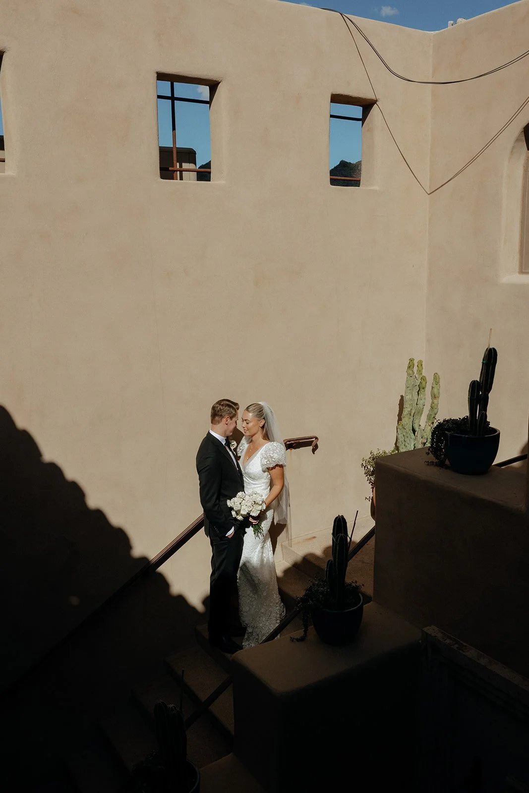 A bride in a white gown holding a bouquet stands next to a groom in a black suit and bow tie in a corridor with tan walls and wooden beams for a destination wedding in Italy