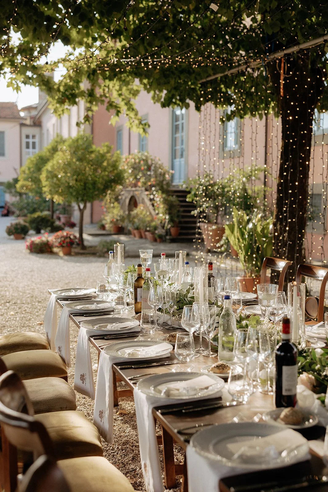 A long outdoor dining table with wooden chairs, set for a meal under trees decorated with string lights and a white runner with greenery for a destination wedding in Italy