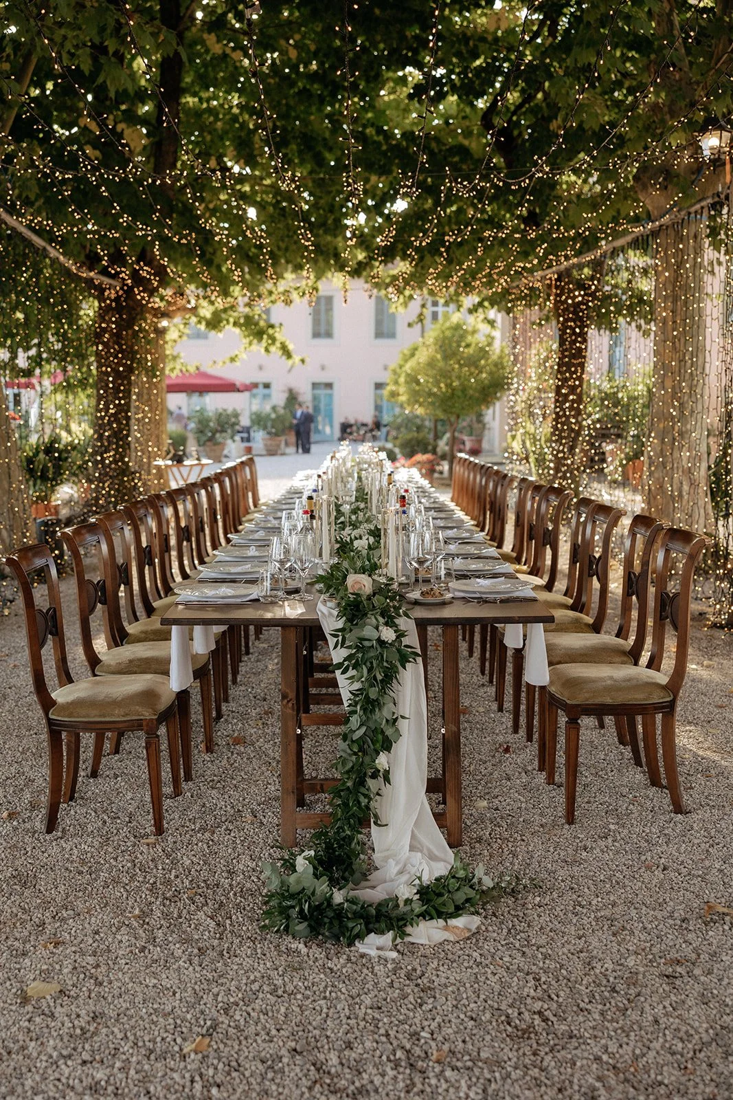 A long outdoor dining table with wooden chairs, set for a meal under trees decorated with string lights and a white runner with greenery for a destination wedding in Italy