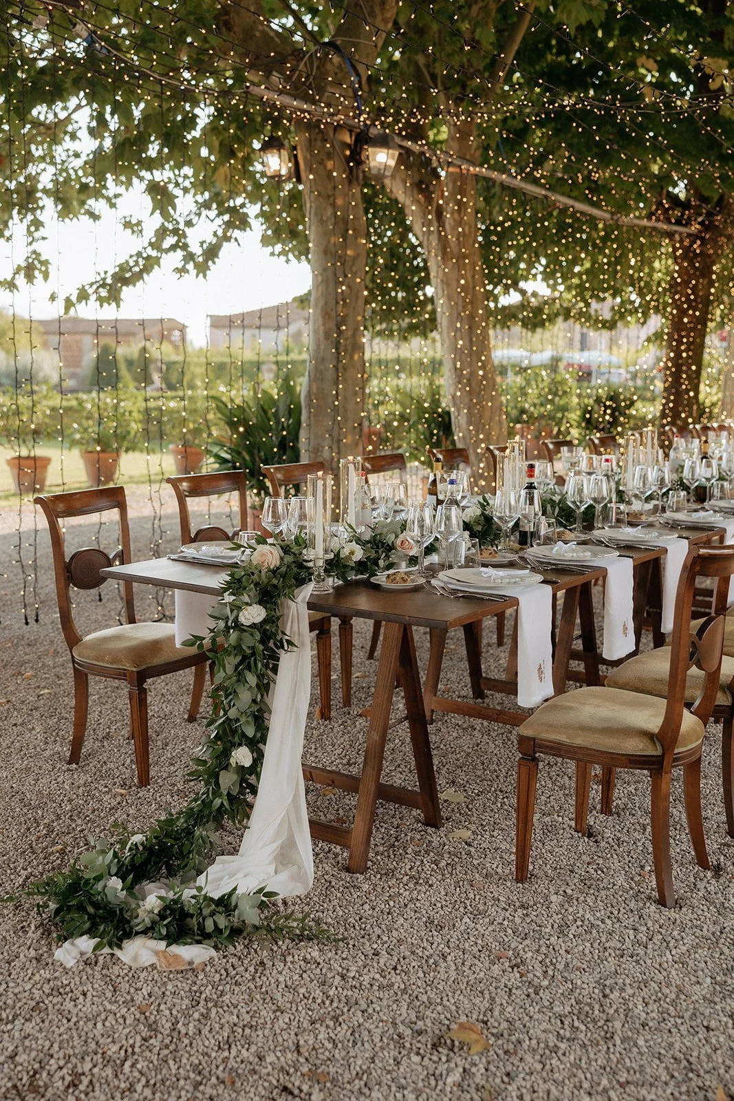 A long outdoor dining table with wooden chairs, set for a meal under trees decorated with string lights and a white runner with greenery for a destination wedding in Italy