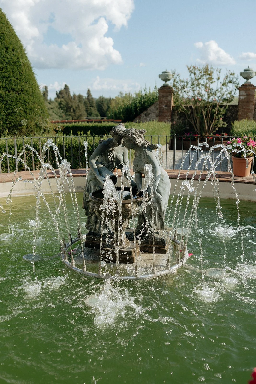 A water fountain with a statue of two figures in the center, surrounded by arched streams of water, in a garden setting.