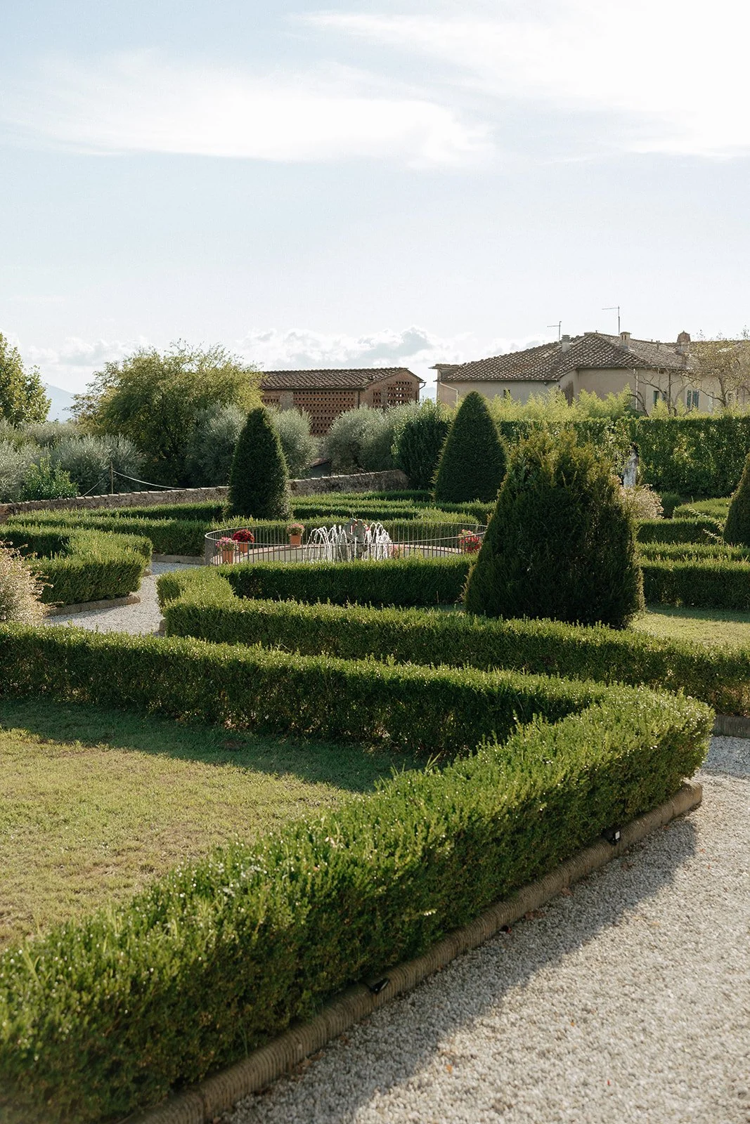 Formal garden with manicured hedges, gravel paths, and topiary bushes, with a building visible in the background under a partly cloudy sky.