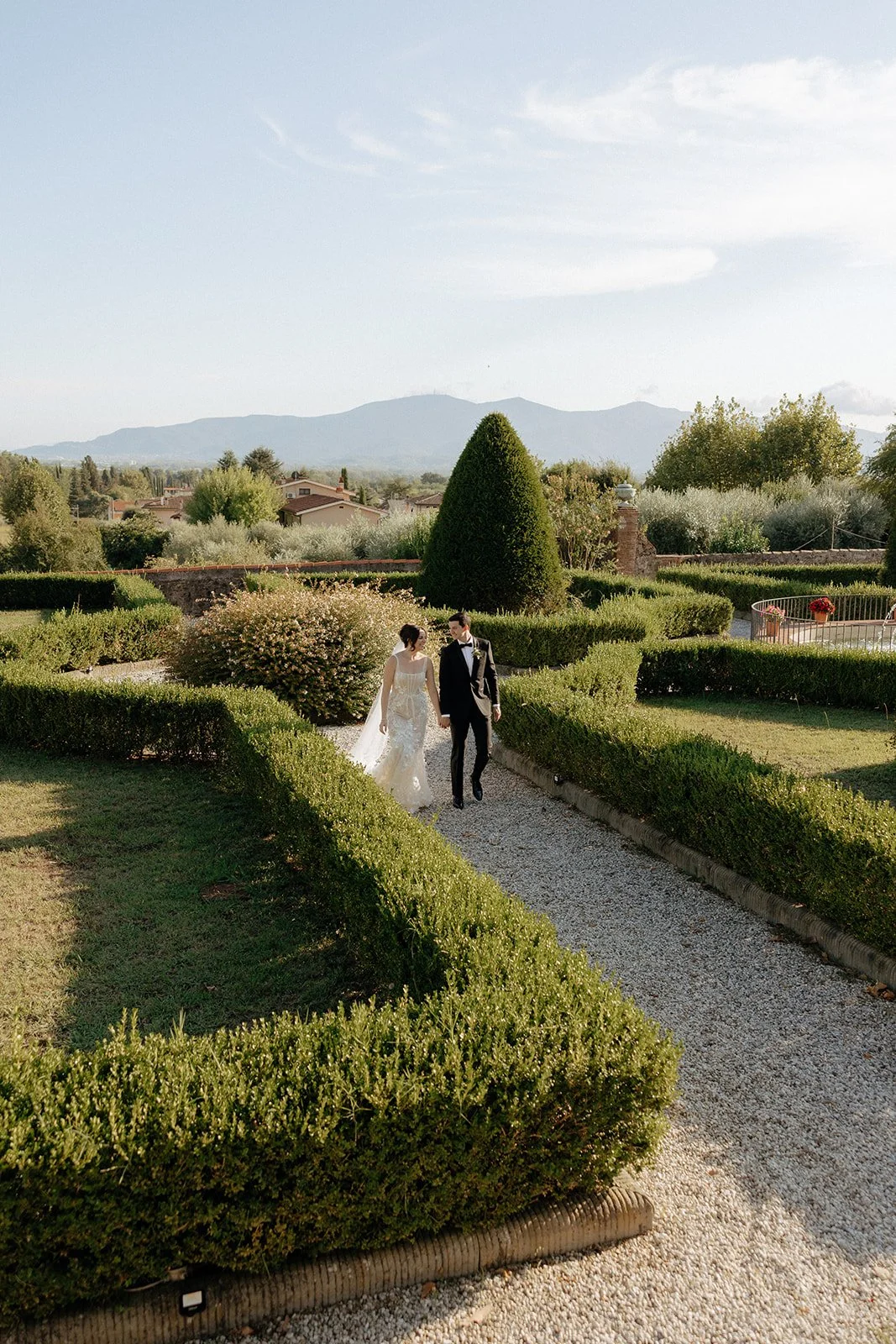 A bride and groom walk along a gravel path through a neatly manicured garden with mountains visible in the background.