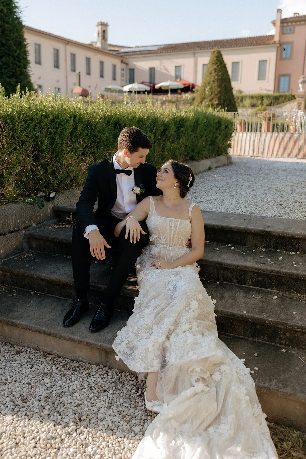A bride and groom in formal wedding attire sit together on outdoor stone steps, looking at each other, with a historic building and greenery in the background.