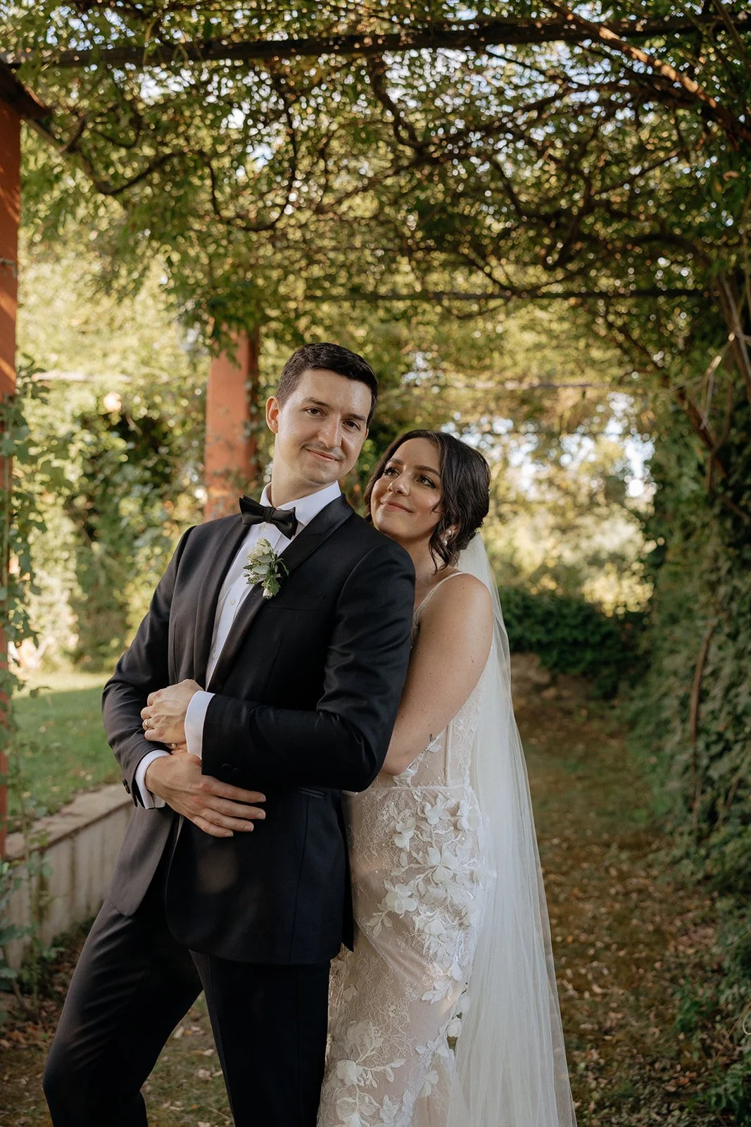 A bride and groom in formal wedding attire sit together on outdoor stone steps, looking at each other, with a historic building and greenery in the background.