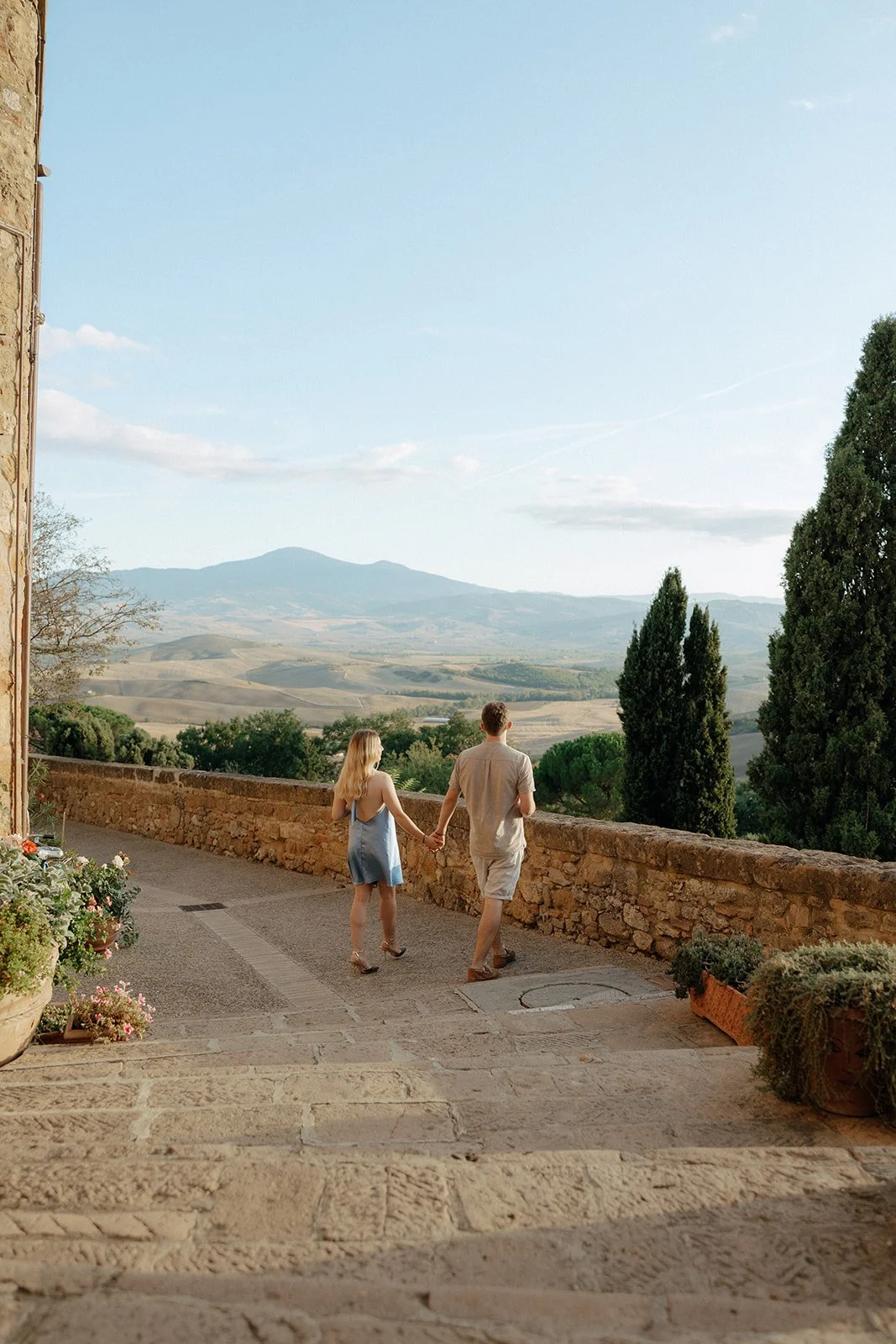 A couple holding hands walks along a stone path bordered by a low wall, with trees and distant hills visible under a clear sky.