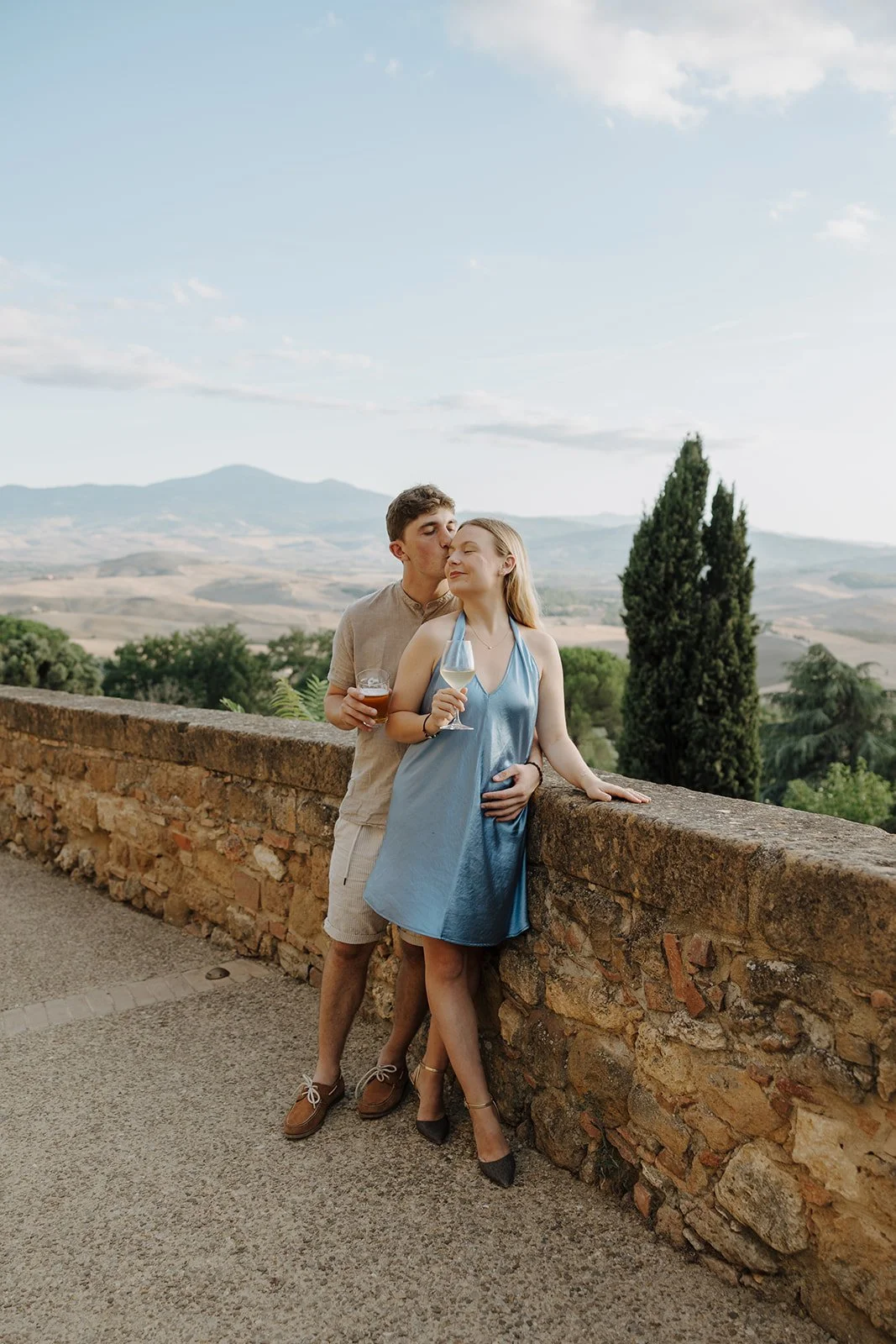 A couple holding hands walks along a stone path bordered by a low wall, with trees and distant hills visible under a clear sky.