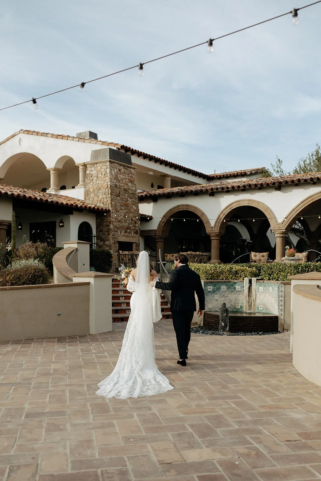 A bride in a white dress and veil stands beside a vintage convertible car while a groom in a suit sits in the driver’s seat outside a stone and stucco building for italy destination wedding