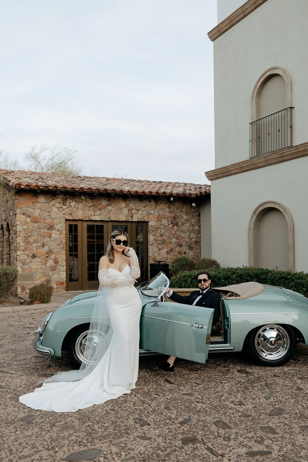 A bride in a white dress and veil stands beside a vintage convertible car while a groom in a suit sits in the driver’s seat outside a stone and stucco building for italy destination wedding