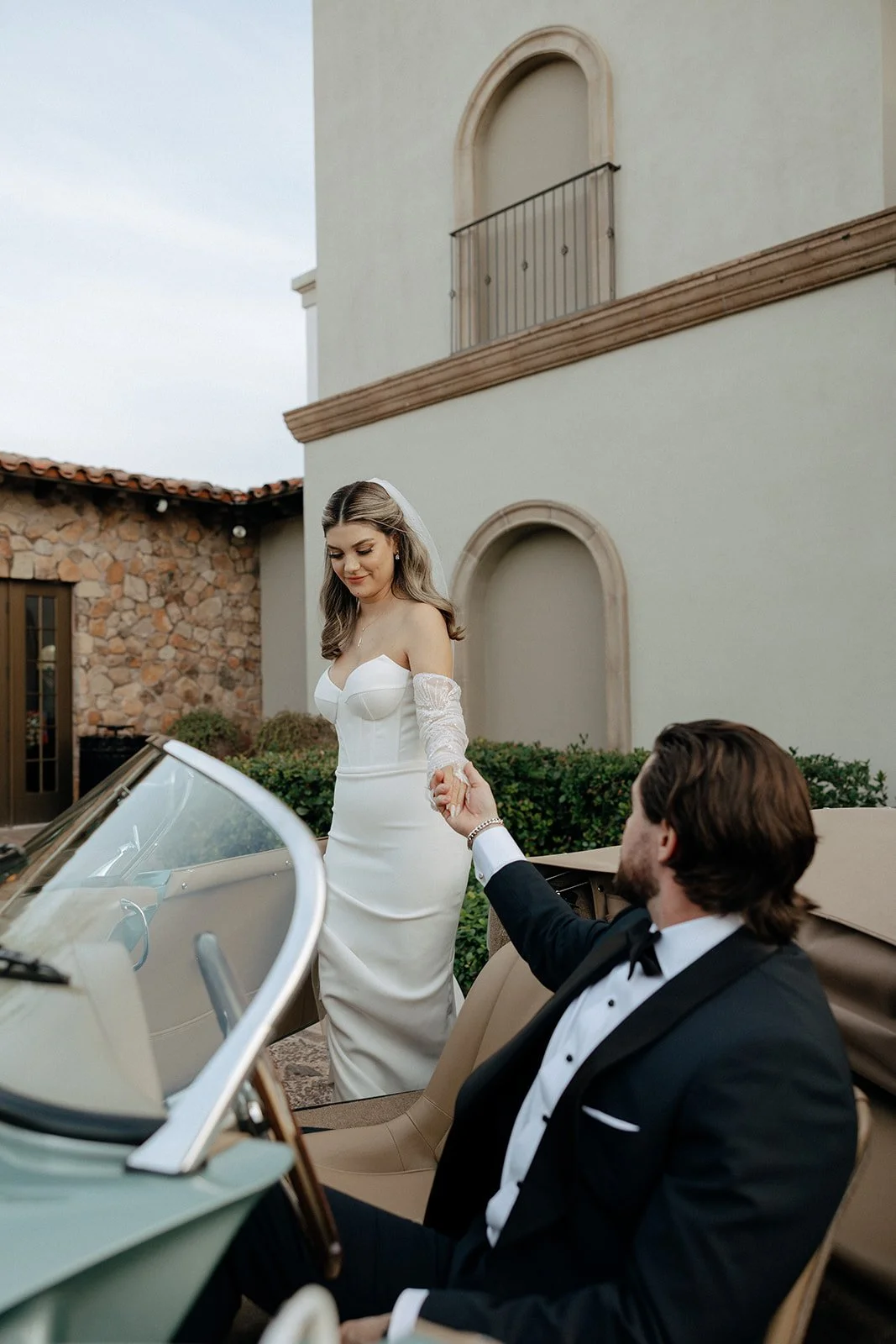A bride in a white dress and veil stands beside a vintage convertible car while a groom in a suit sits in the driver’s seat outside a stone and stucco building for italy destination wedding