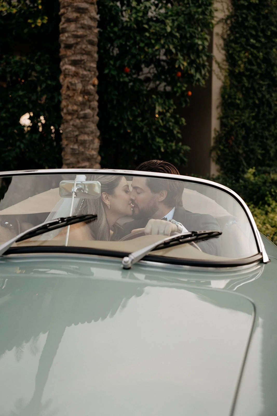 A bride in a white dress and veil stands beside a vintage convertible car while a groom in a suit sits in the driver’s seat outside a stone and stucco building for italy destination wedding