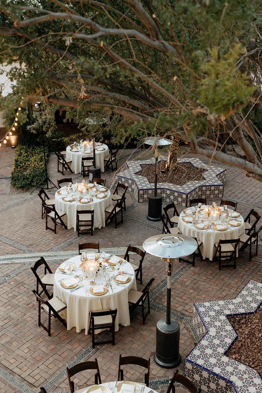 Outdoor courtyard with round tables set for an event, white tablecloths, wooden chairs, and candlelit centerpieces under large trees for an Italy destination wedding 