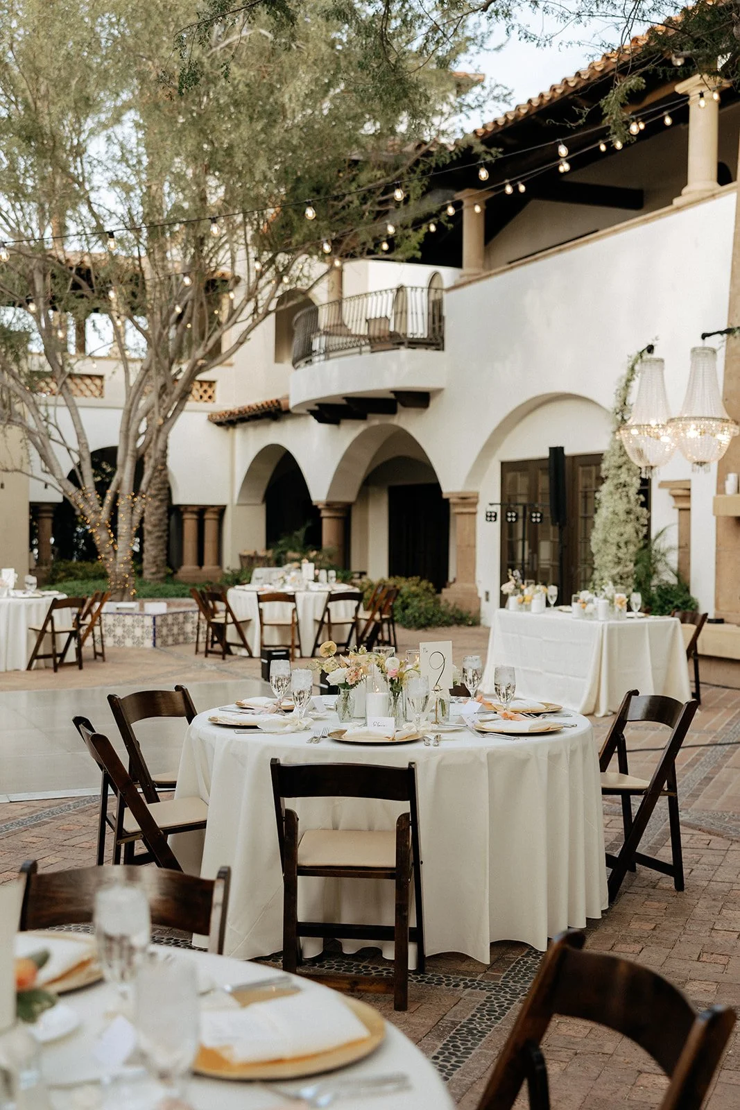 Outdoor courtyard with round tables set for an event, white tablecloths, wooden chairs, and candlelit centerpieces under large trees for an Italy destination wedding 