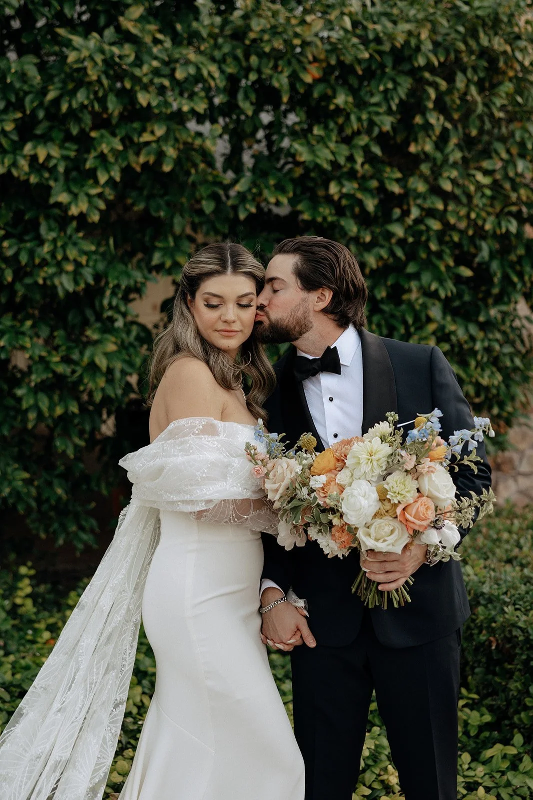 A bride and groom stand outdoors facing each other, the bride holding a bouquet and wearing a long white gown, with greenery and a stone wall in the background.