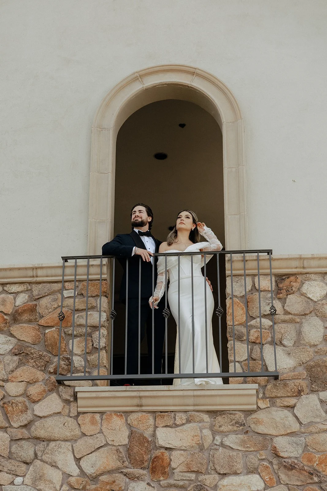A bride and groom stand outdoors facing each other, the bride holding a bouquet and wearing a long white gown, with greenery and a stone wall in the background.