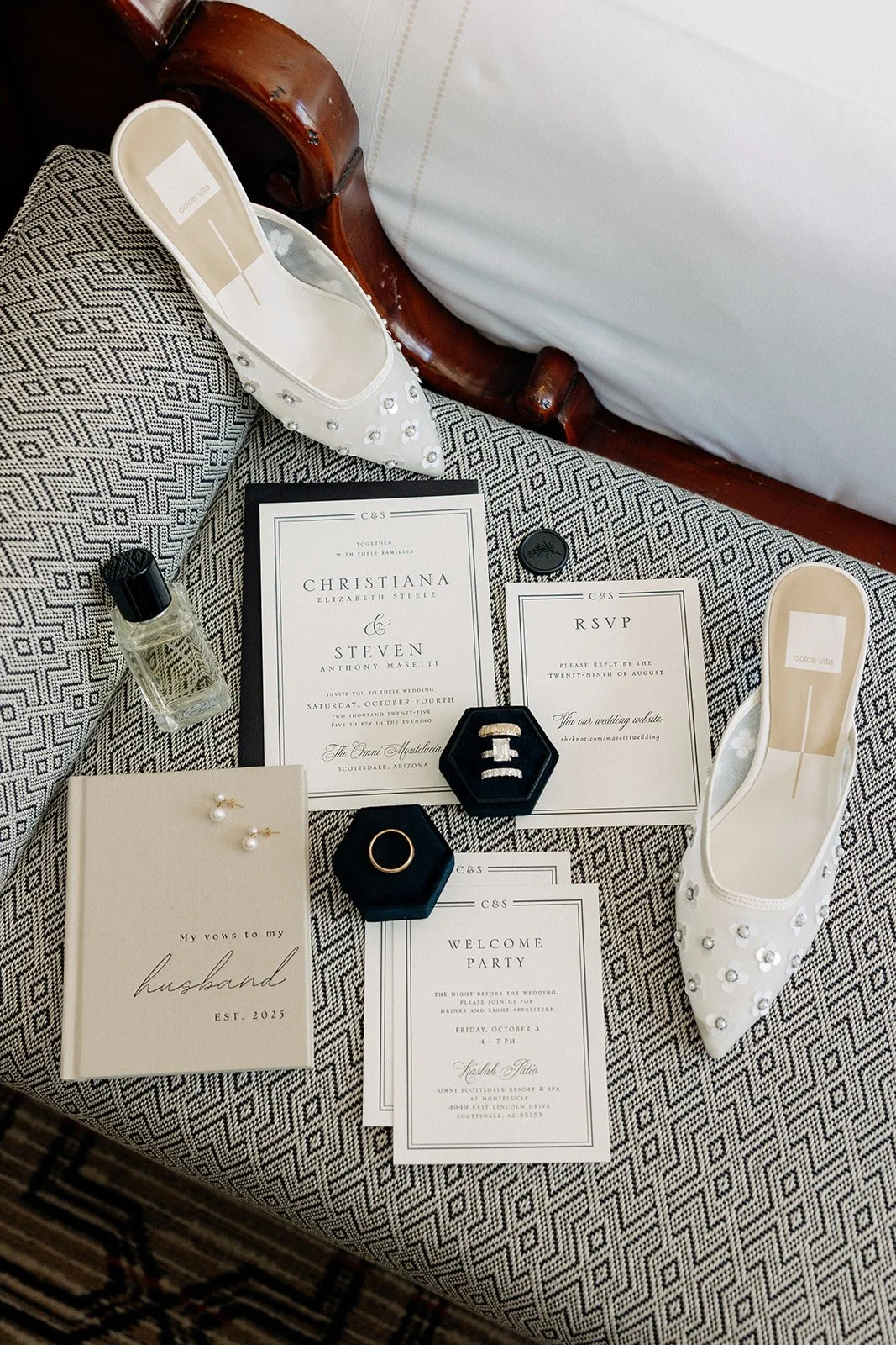 Flat lay of wedding items including jeweled white shoes, invitation cards, a perfume bottle, two ring boxes, a notebook, and pearl earrings on a patterned bench.