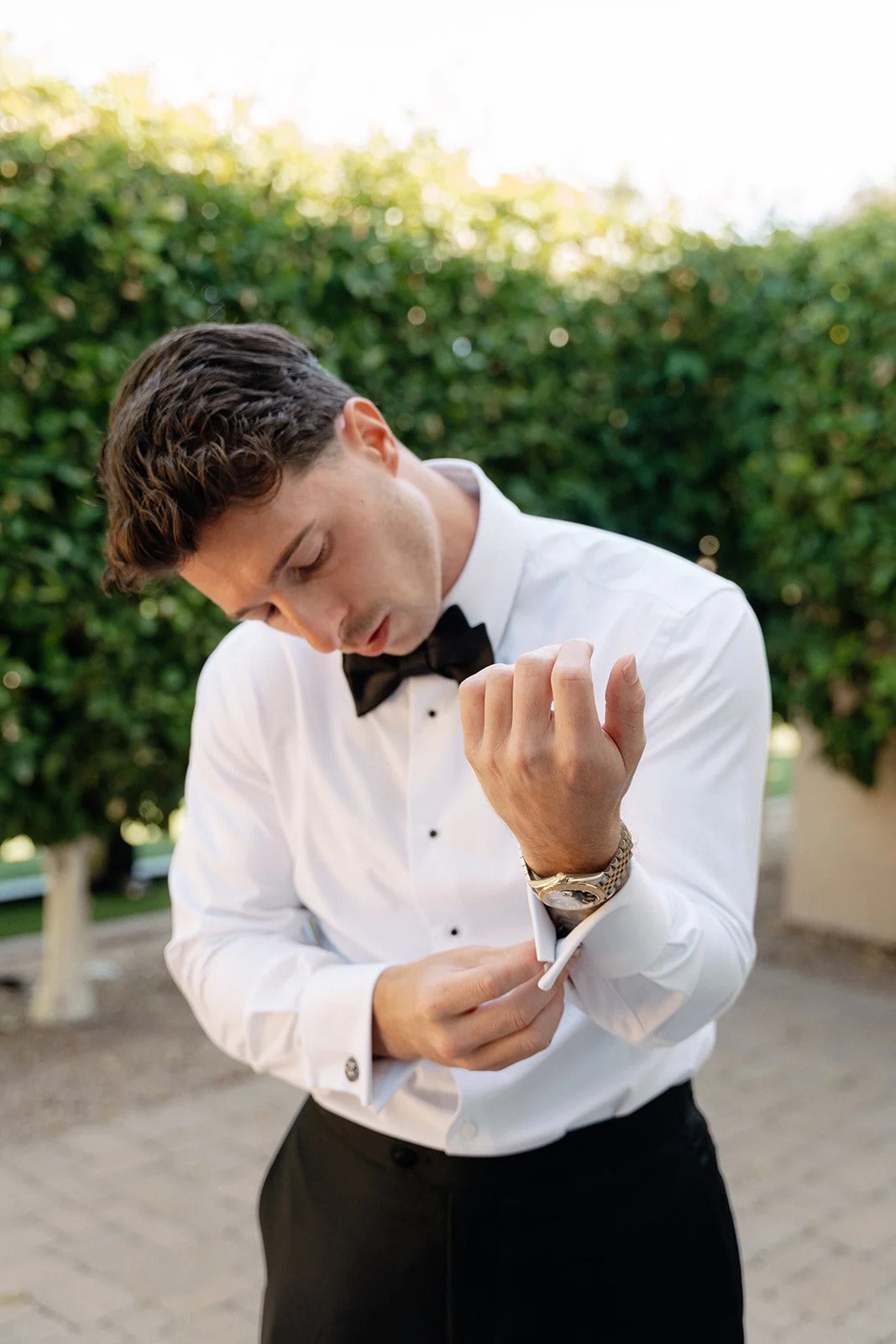 A man in a white dress shirt and black bow tie fastens his cufflink outdoors, standing on a paved area with green bushes in the background.