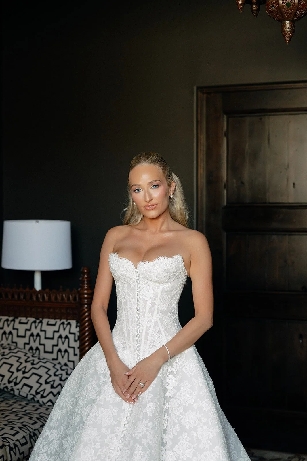 A woman in a white strapless lace wedding dress stands indoors with her hands clasped, in front of a patterned chair and a dark wooden door for a luxury wedding