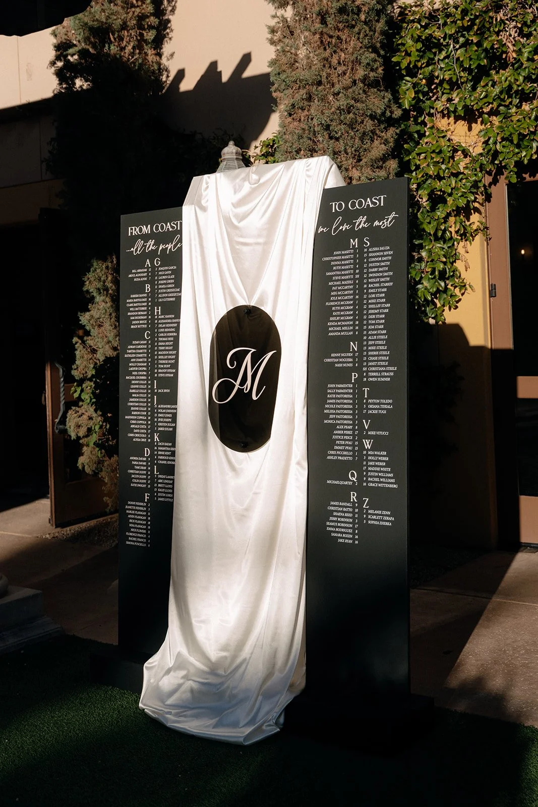 Two black boards with guest names listed alphabetically are partially covered by a white draped cloth with a large "M" in the center, standing outdoors near greenery.