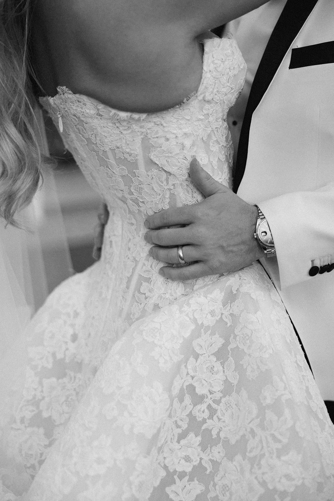 A close-up, black and white photo of a bride and groom embracing; the bride wears a lace wedding dress and the groom wears a light suit with a watch and wedding ring.