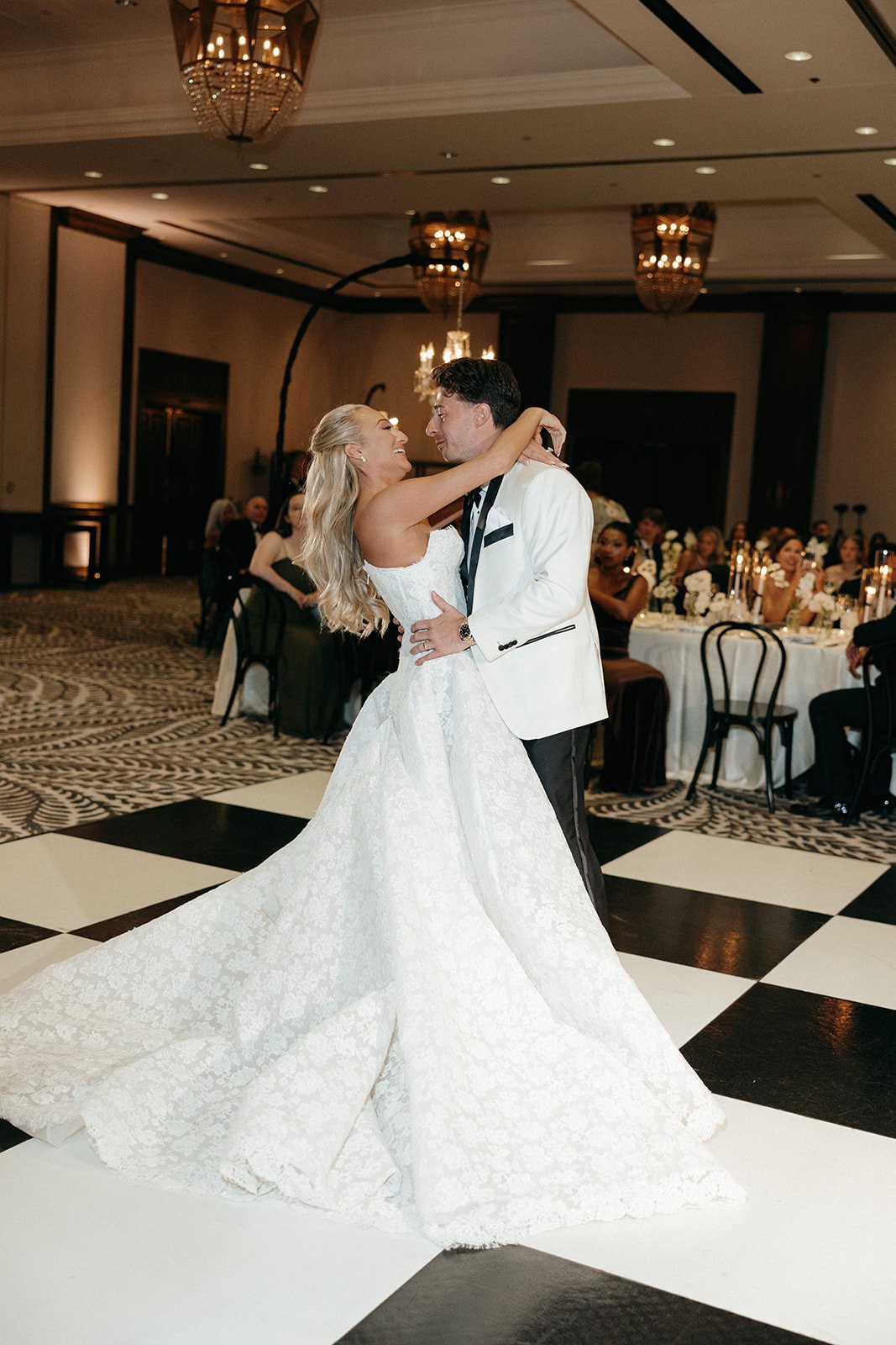 A bride and groom dance together on a black and white checkered floor in a formal, decorated ballroom with seated guests watching.