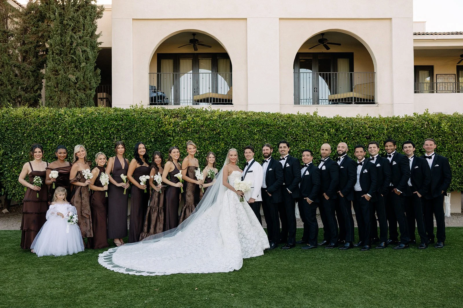A bride in a white gown stands with eleven bridesmaids wearing dark brown dresses, all holding bouquets, posing outside in front of a large building and greenery.