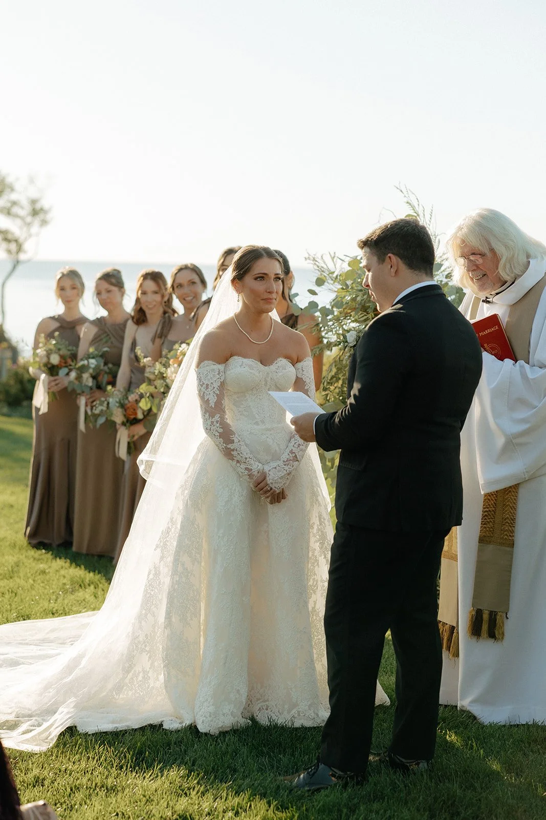 A bride and groom stand with an officiant at an outdoor wedding ceremony, surrounded by guests seated on either side, with blue sky and greenery in the background for a wedding in new york