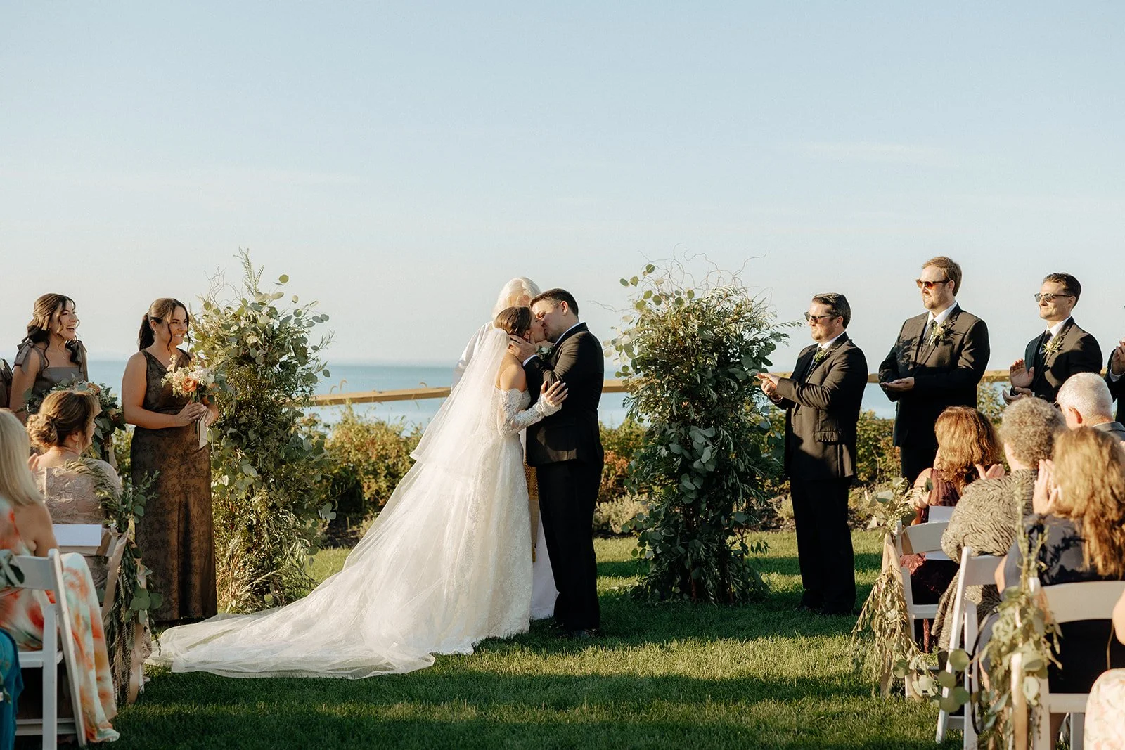 A bride and groom stand with an officiant at an outdoor wedding ceremony, surrounded by guests seated on either side, with blue sky and greenery in the background for a wedding in new york