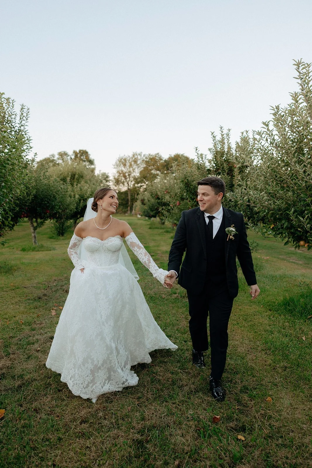 A bride and groom stand together in an orchard, dressed in wedding attire, with trees and grass surrounding them for a wedding in new york