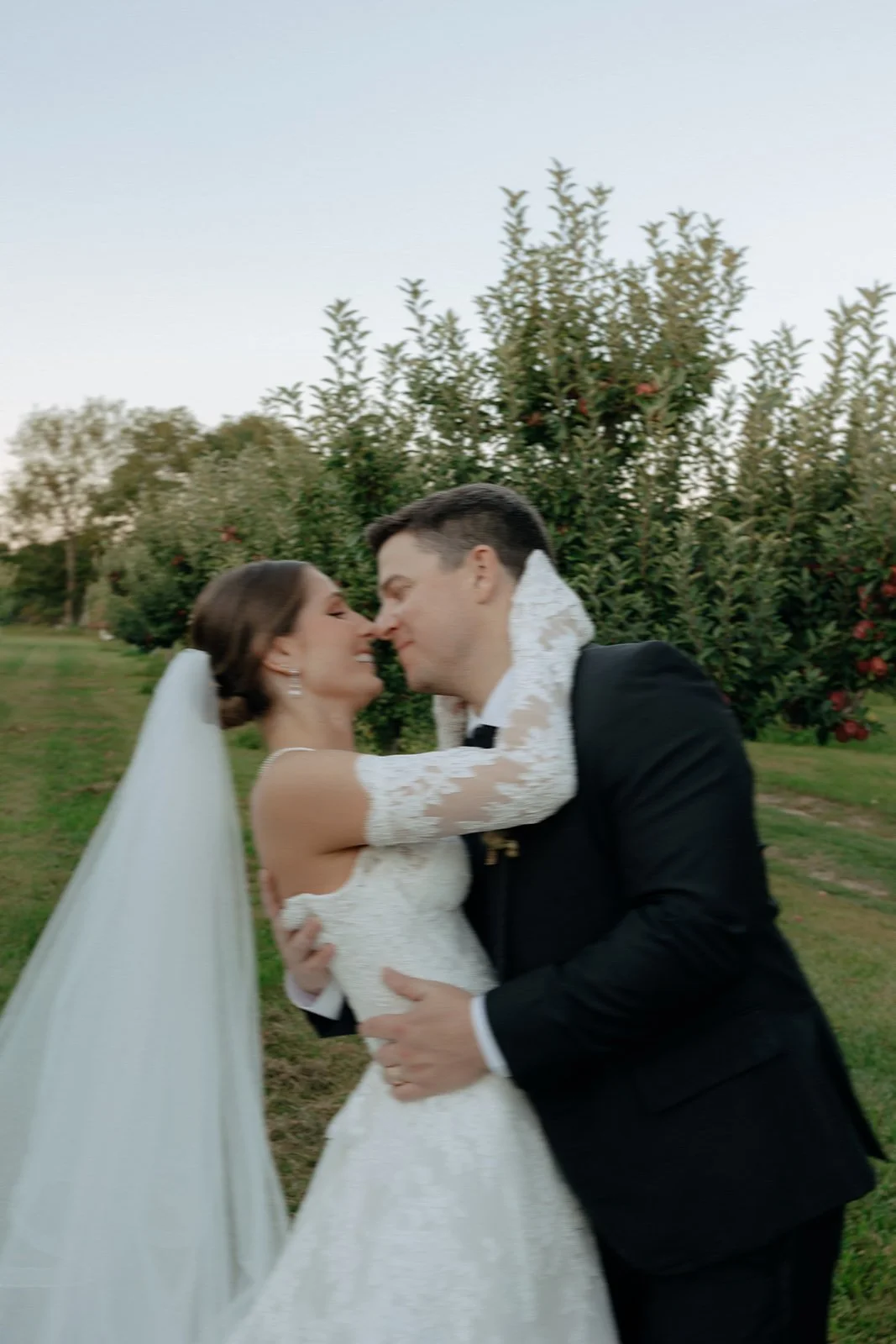 A bride and groom stand together in an orchard, dressed in wedding attire, with trees and grass surrounding them for a wedding in new york