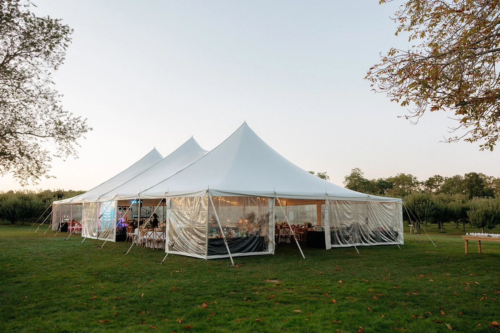 Large white event tent with clear side panels set up on a grassy lawn, with people and tables visible inside, surrounded by trees under a clear sky.