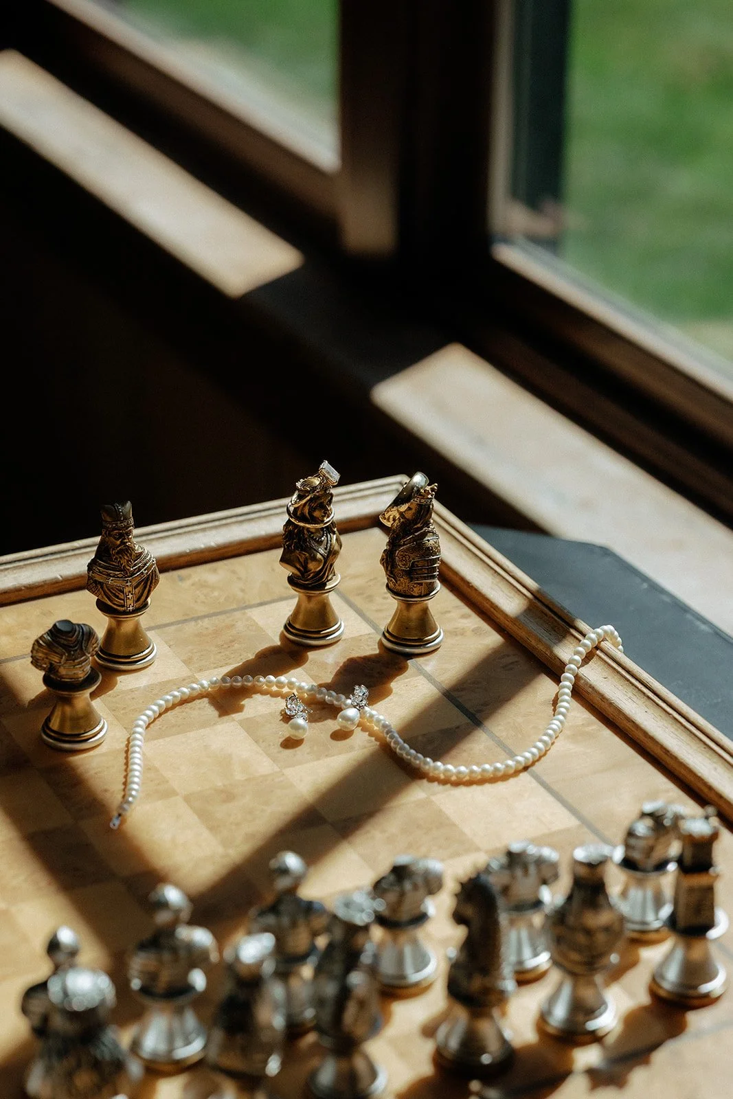 A pearl necklace and earrings rest on the edge of a decorative chessboard near a window with sunlight streaming in.