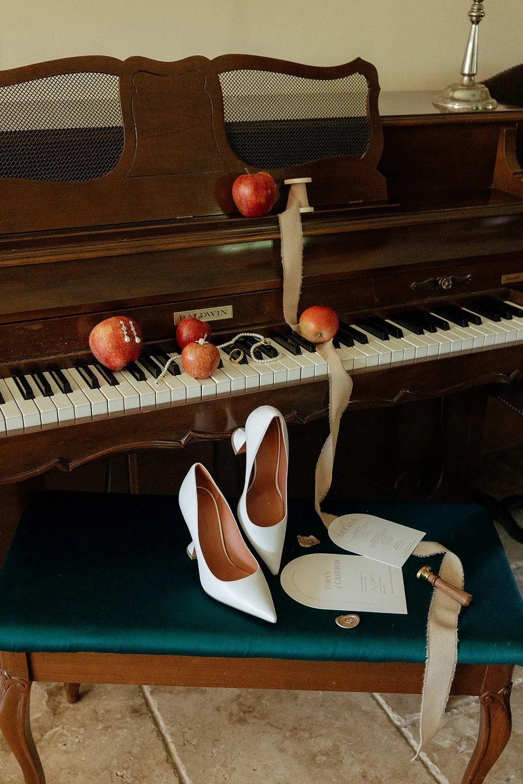 A brown piano with apples on the keys, a pair of white high-heeled shoes on the bench, and papers with ribbon placed nearby.