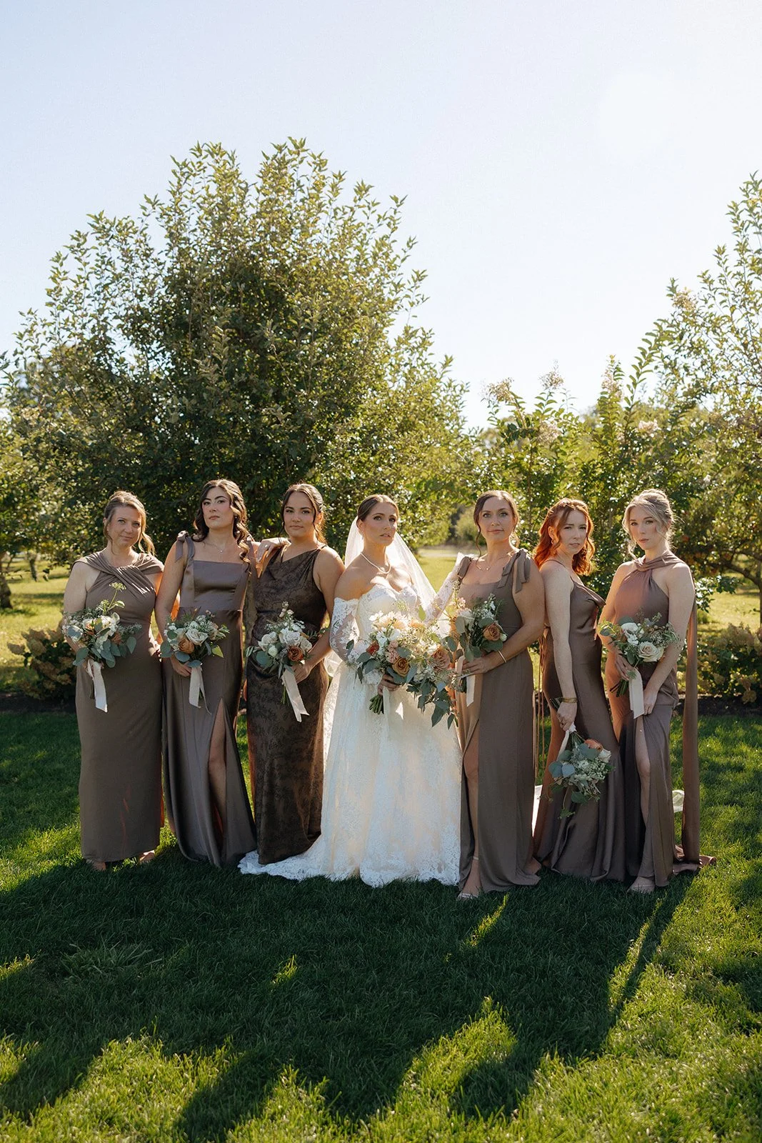 A bride in a white dress stands outdoors with six bridesmaids in brown dresses, all holding bouquets, in front of green trees on a sunny day.