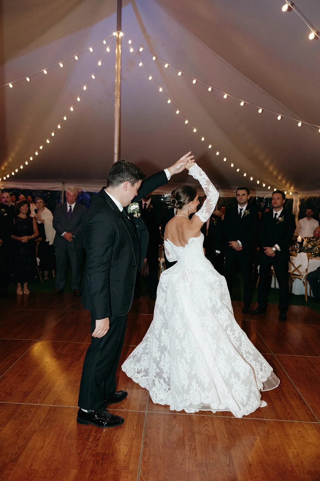 A bride and groom share a dance on a wooden floor under string lights at their wedding reception, with guests watching in the background.