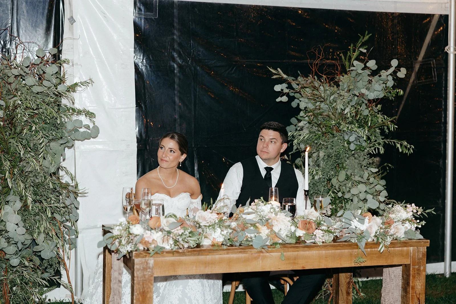 A woman in a formal dress hands a gift box to a bride seated at a decorated table, while others look on at a wedding reception.