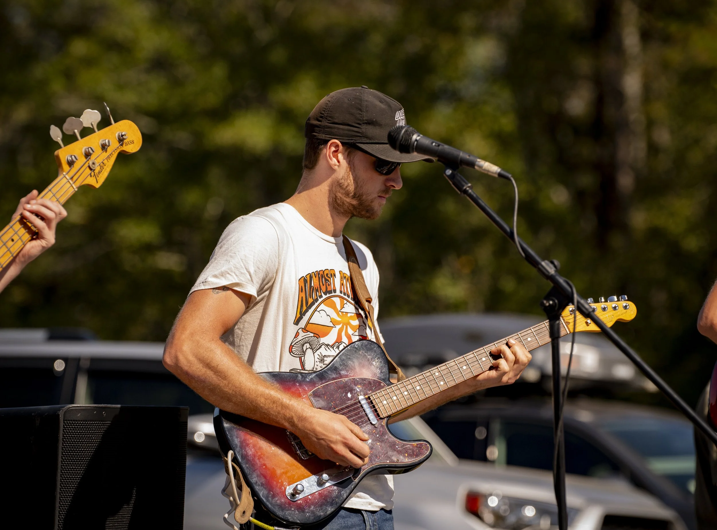 A man wearing sunglasses, a black cap, and a white T-shirt playing an electric guitar during an outdoor music performance.