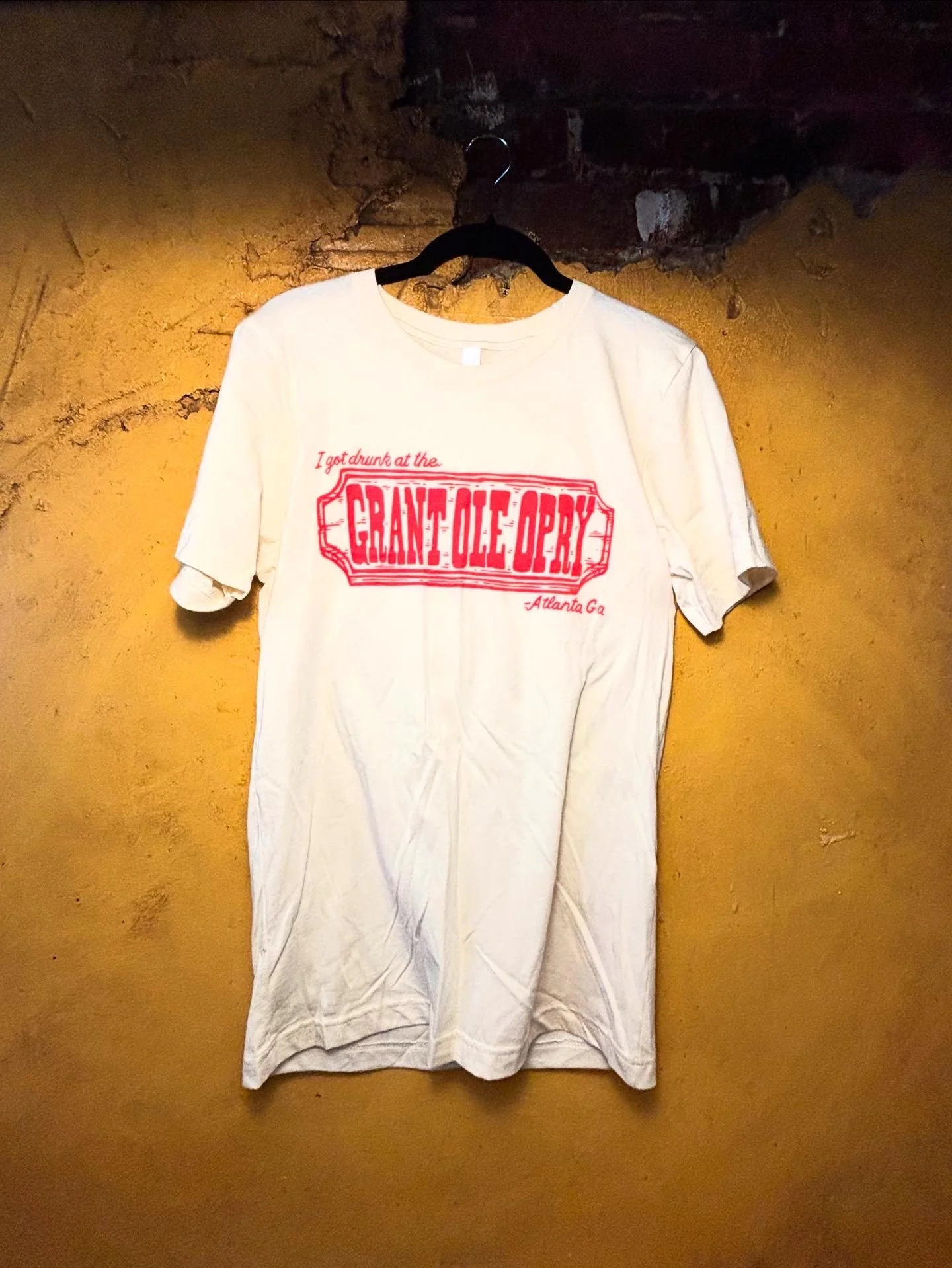 White t-shirt with red text that reads, 'I got drunk at the Grant Ole Opry, Atlanta GA,' hanging on a black hanger against a yellow wall.