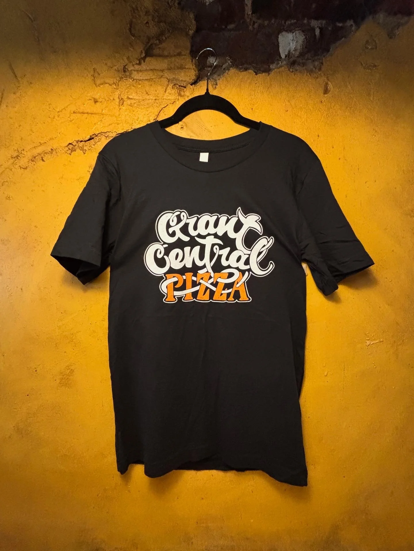 Black T-shirt hanging on a hook with the text "Ground Control Pizza" in white and orange lettering.
