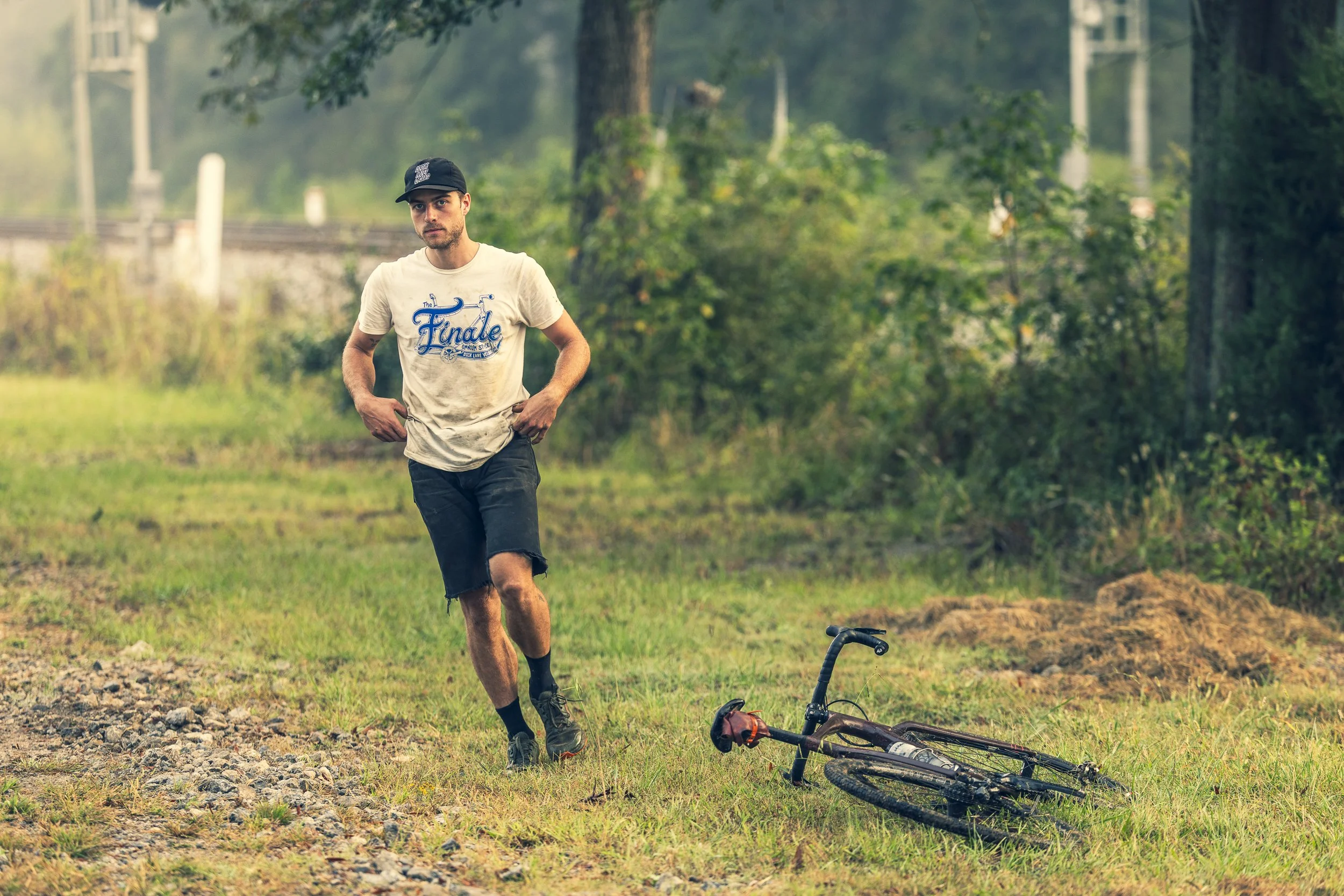 A young man in a white t-shirt with the word 'Finale' on it, black shorts, and a black cap, stands in a grassy area near a bicycle lying on the ground, with trees and a power line in the background.